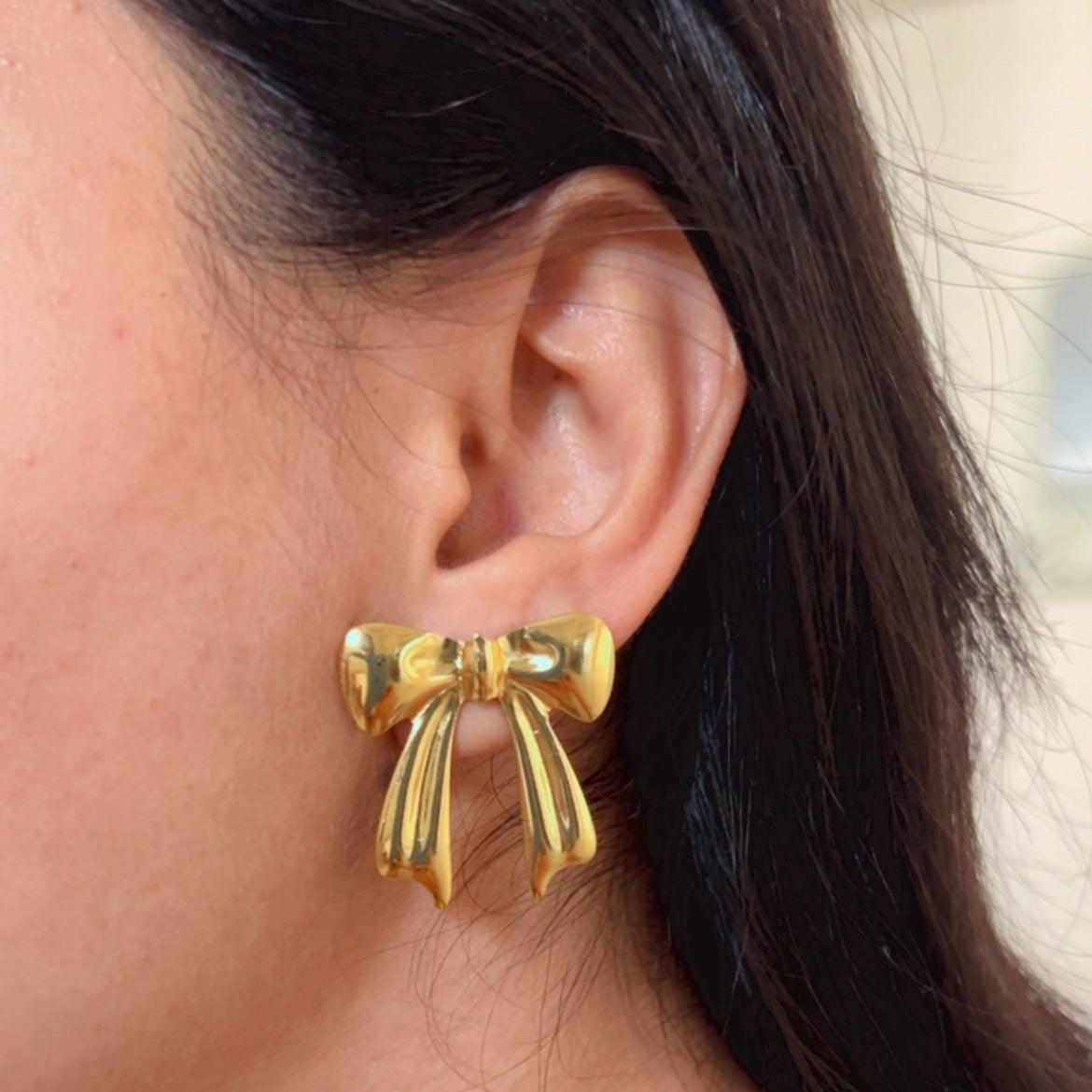 Gold ribbon bow shaped stud earrings on pastel pink surface.