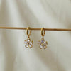 Gold hoop earrings with hanging flower-shaped crystal charms displayed on a wooden rod.