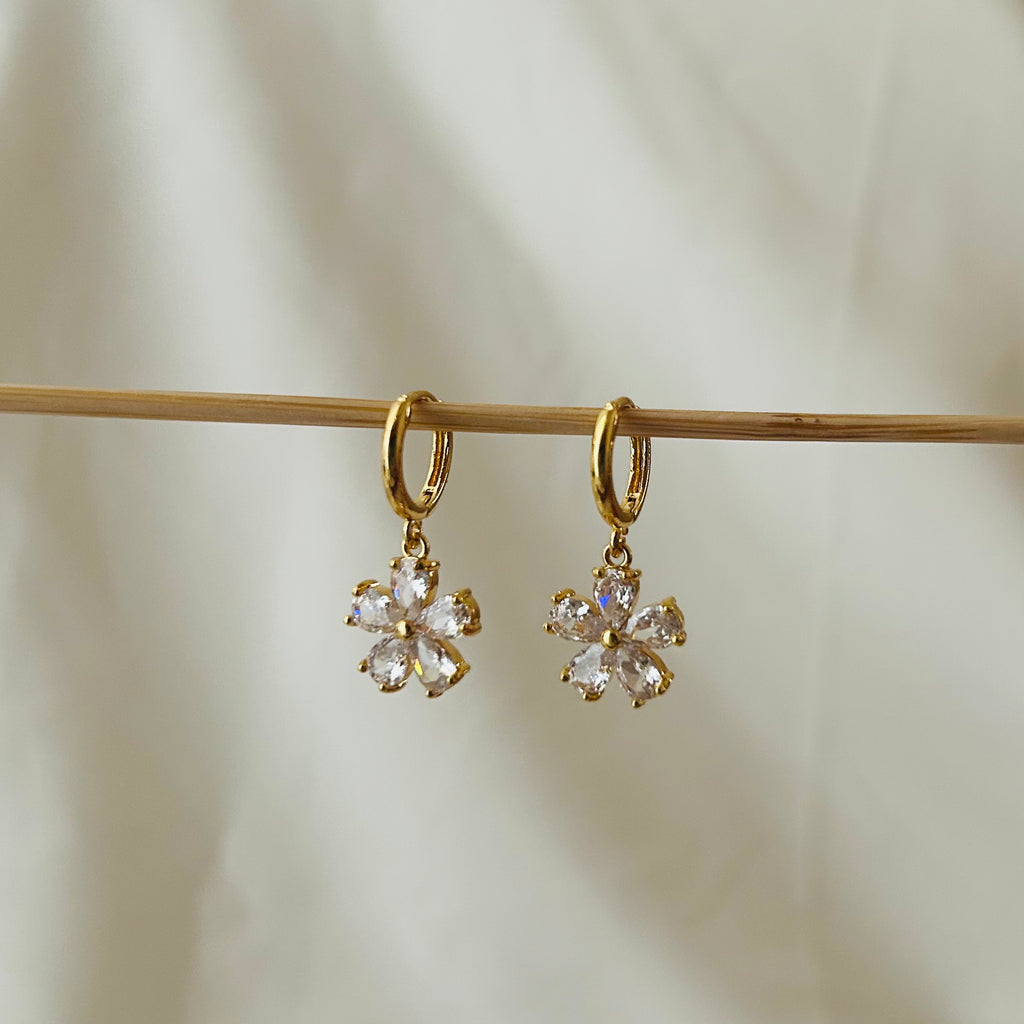 Gold hoop earrings with hanging flower-shaped crystal charms displayed on a wooden rod.