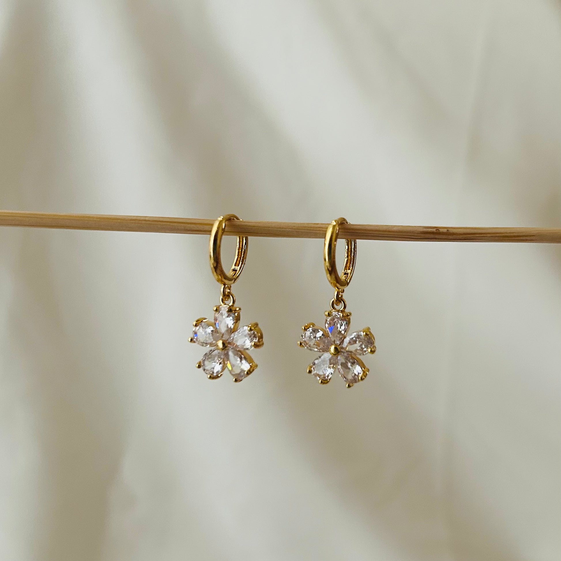Gold hoop earrings with hanging flower-shaped crystal charms displayed on a wooden rod.