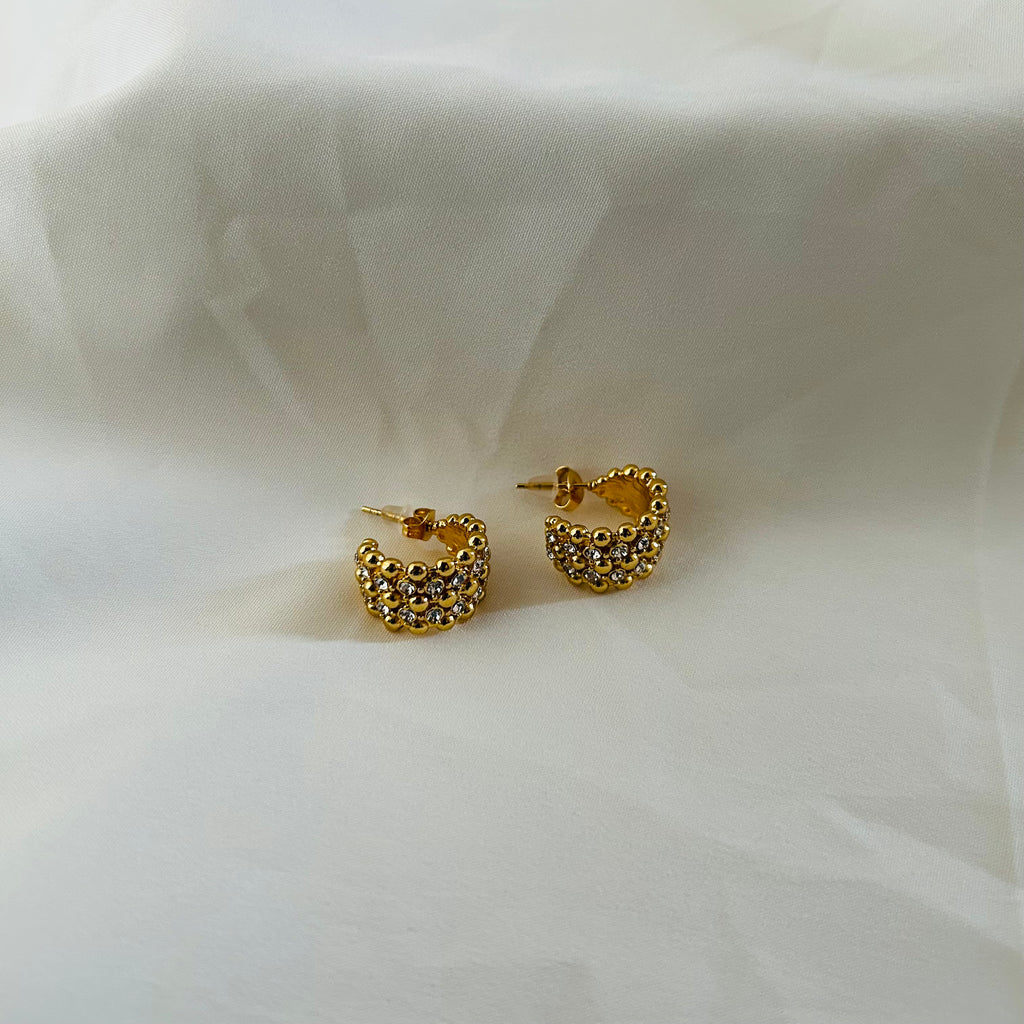 Gold crescent-shaped stud earrings covered with crystal stones on white fabric background.