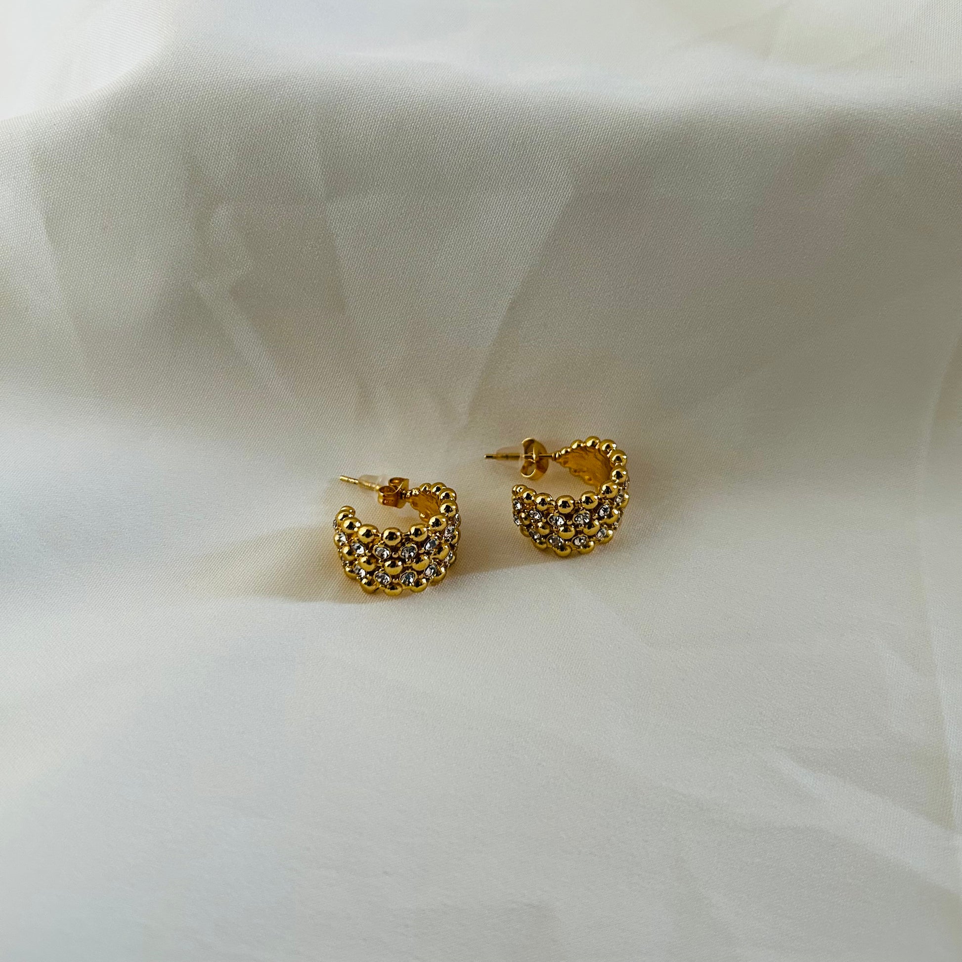 Gold crescent-shaped stud earrings covered with crystal stones on white fabric background.