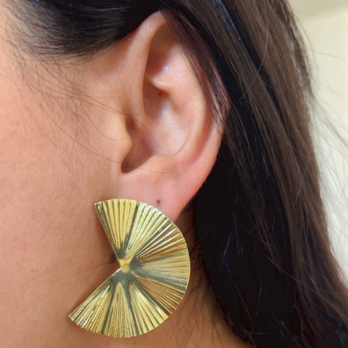 Gold fan shaped textured stud earrings displayed on soft pink surface.