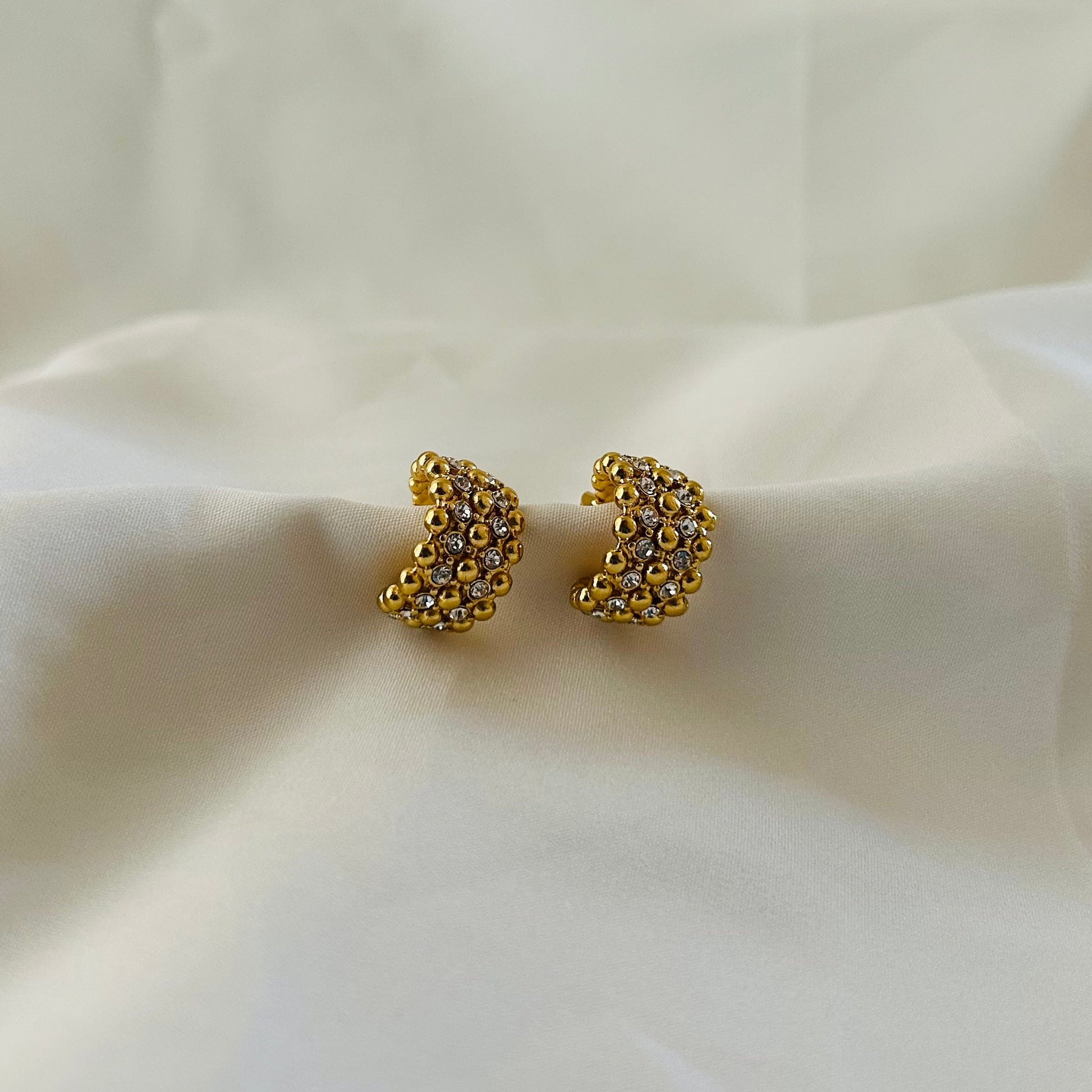Gold crescent-shaped stud earrings covered with crystal stones on white fabric background.