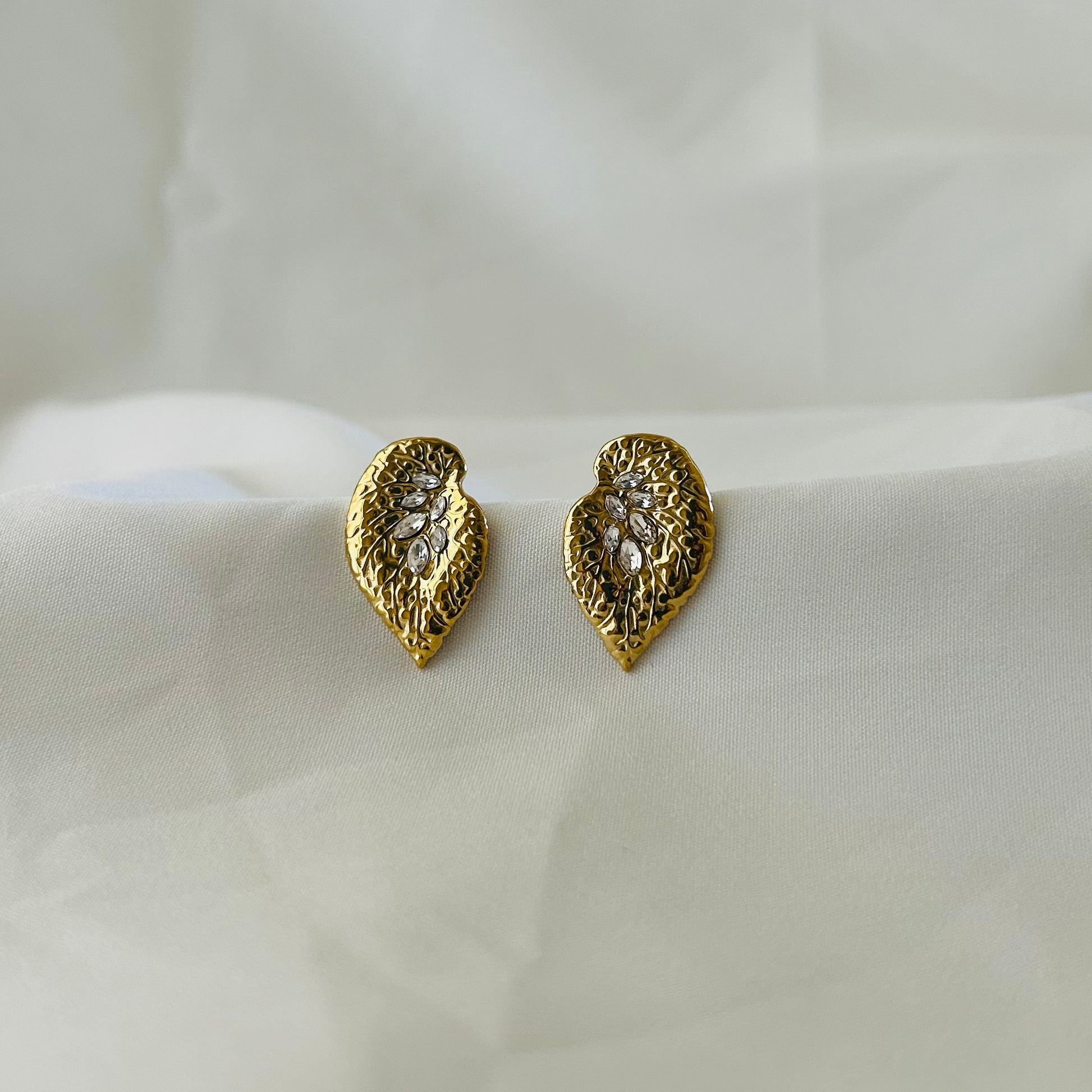 Textured gold leaf-shaped stud earrings with crystal accents displayed on white fabric.