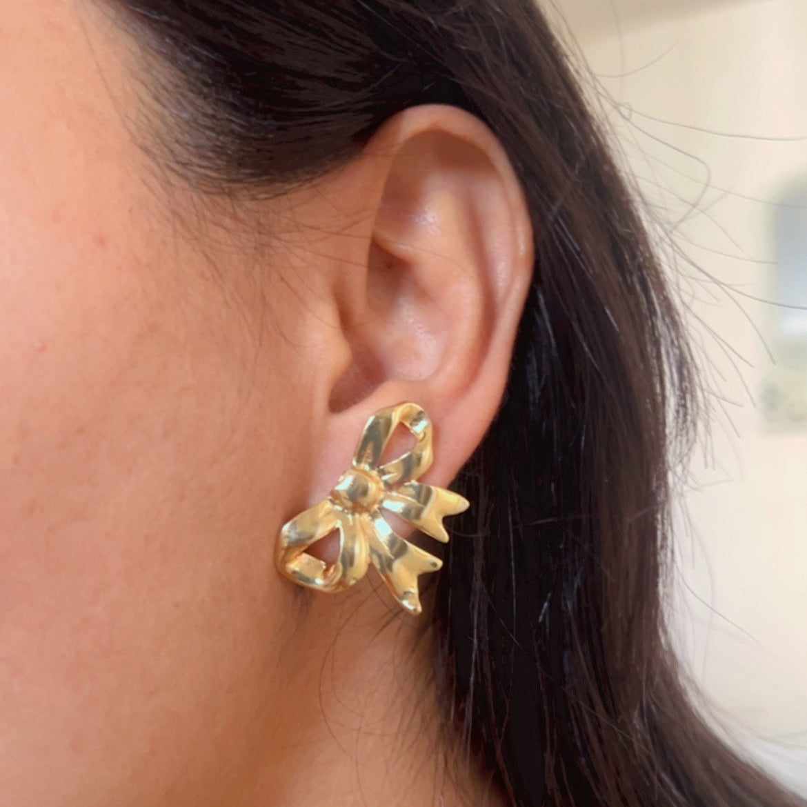 Small gold bow design stud earrings displayed on pink textured background.