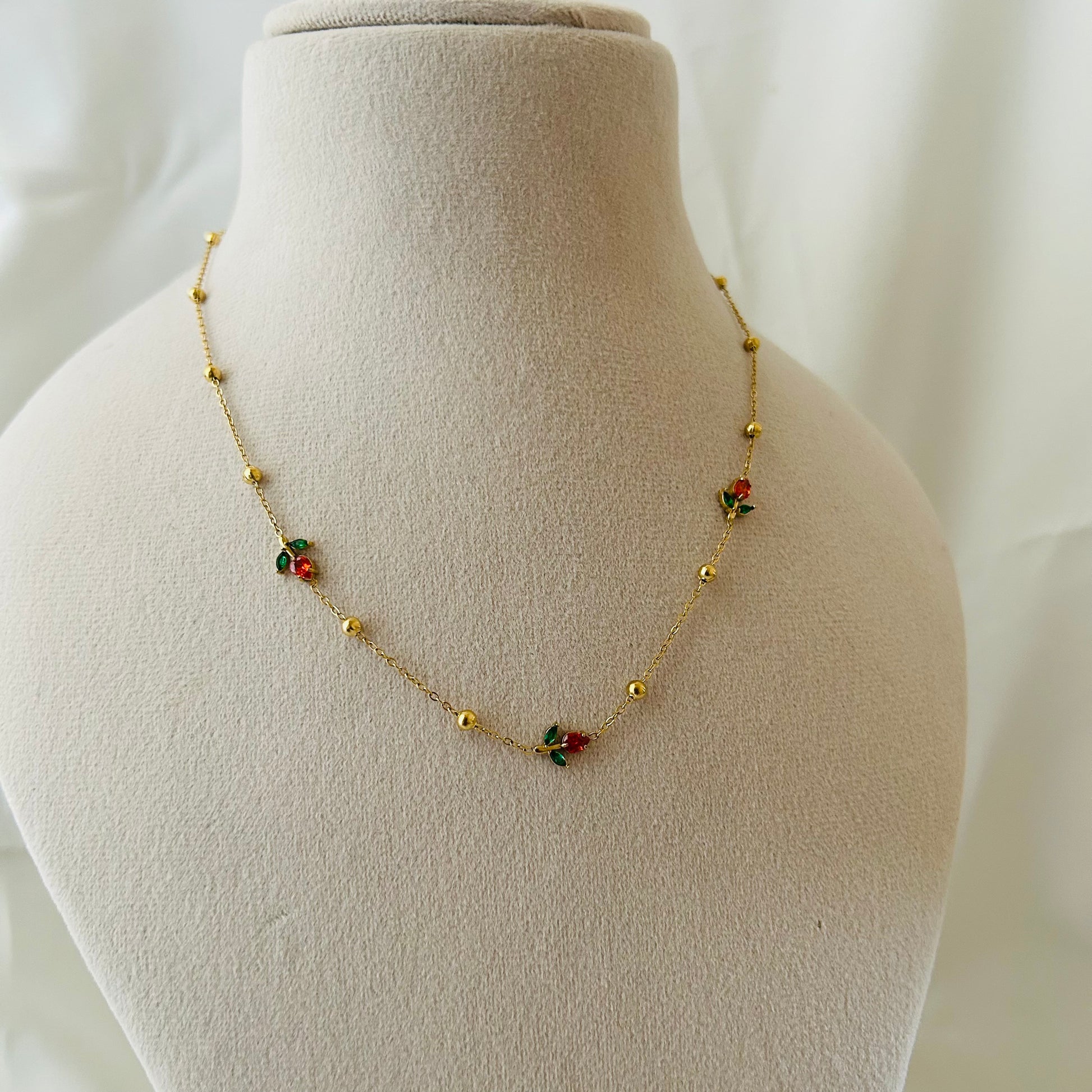 Minimal gold necklace with tiny red and green floral charms displayed on a mannequin bust.