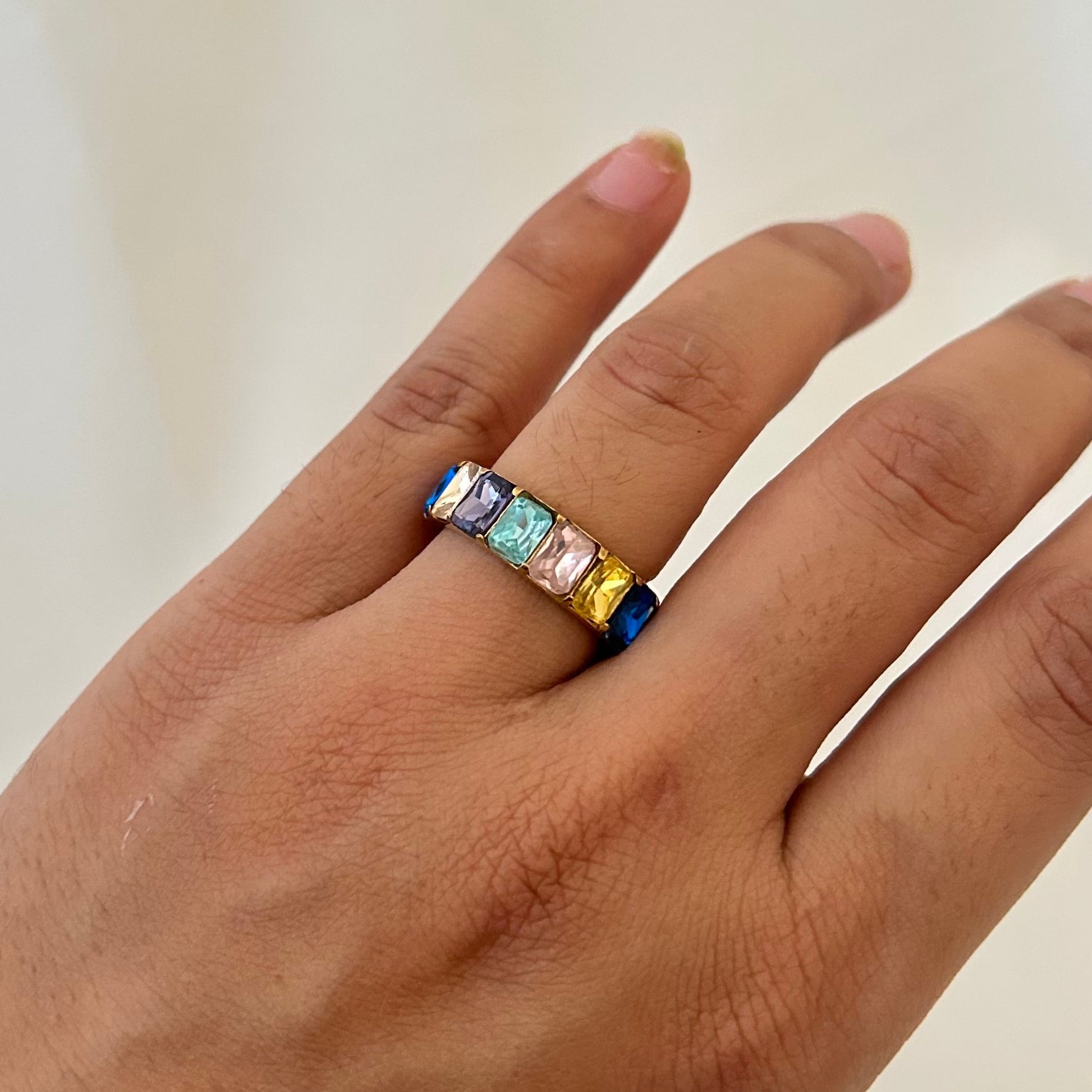 Hand wearing a gold band ring with rectangular multicolor gemstones in a rainbow arrangement.