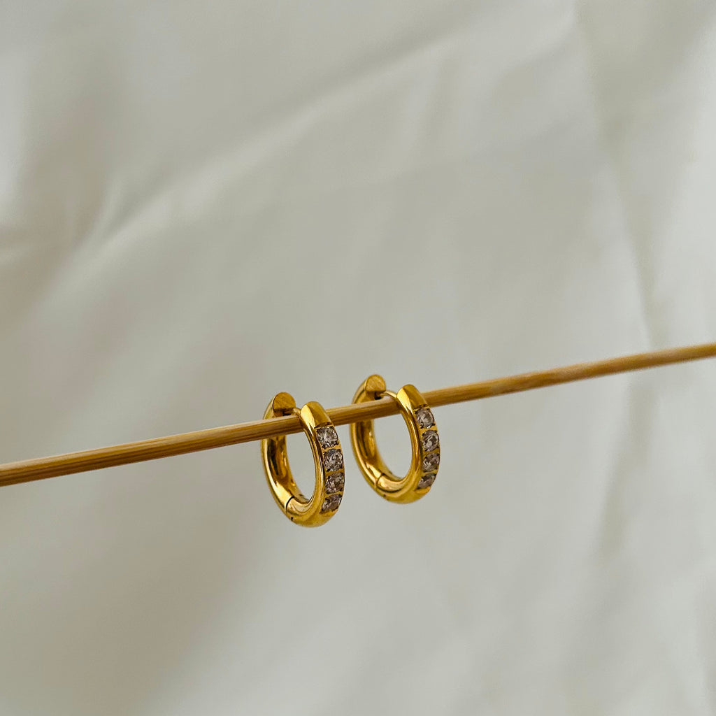 Gold hoop earrings with embedded crystal stones hanging on a wooden display rod.