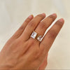 Hand wearing a gold open ring with two clear gemstones, one rectangular and one teardrop-shaped.