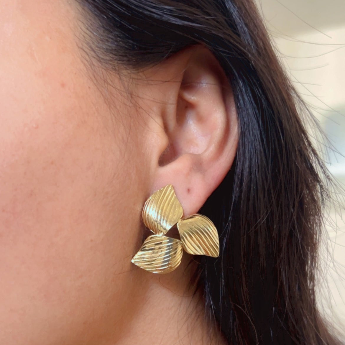 Gold triple leaf textured stud earrings on pink background.