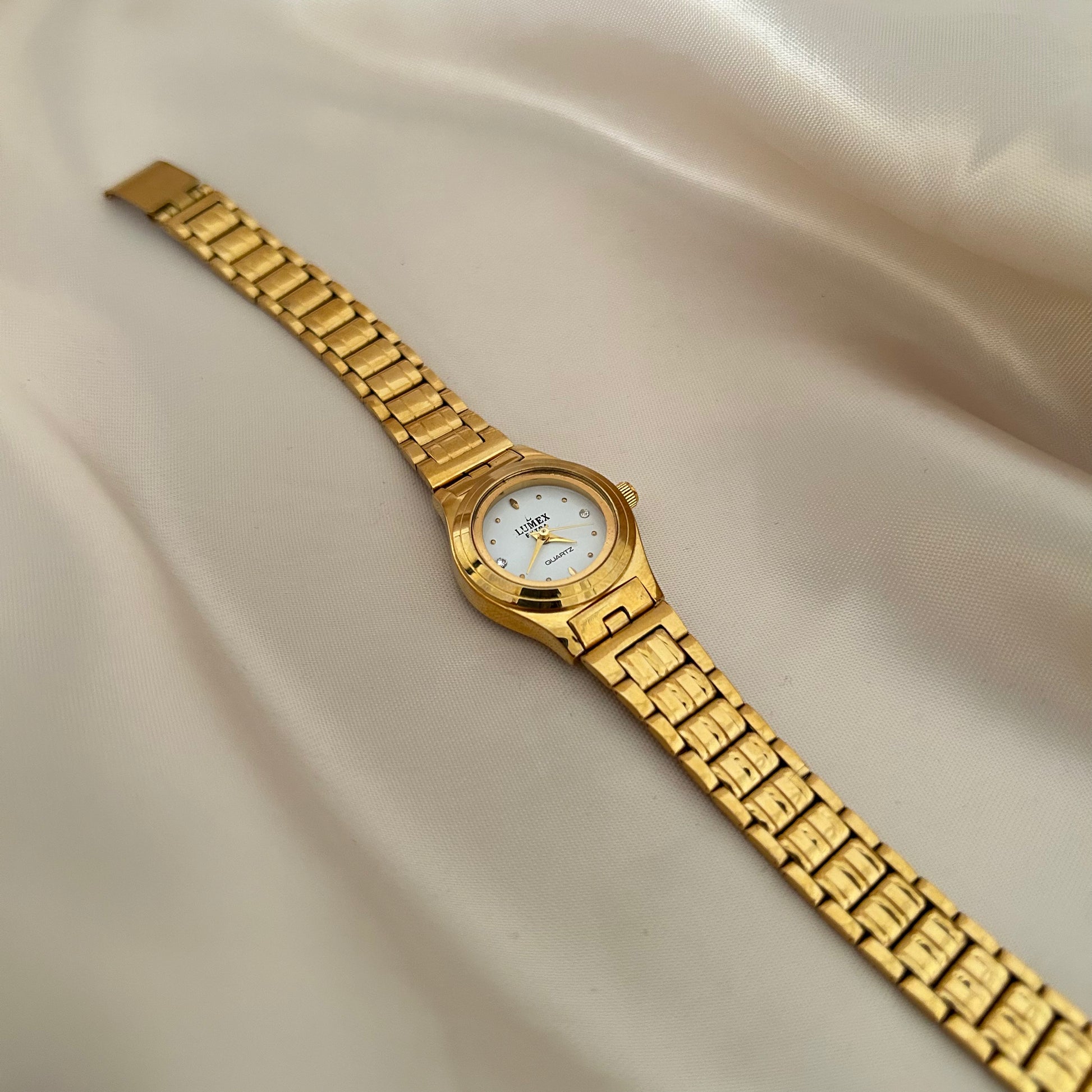 Round gold wristwatch with minimal dial and metallic strap on satin fabric background.