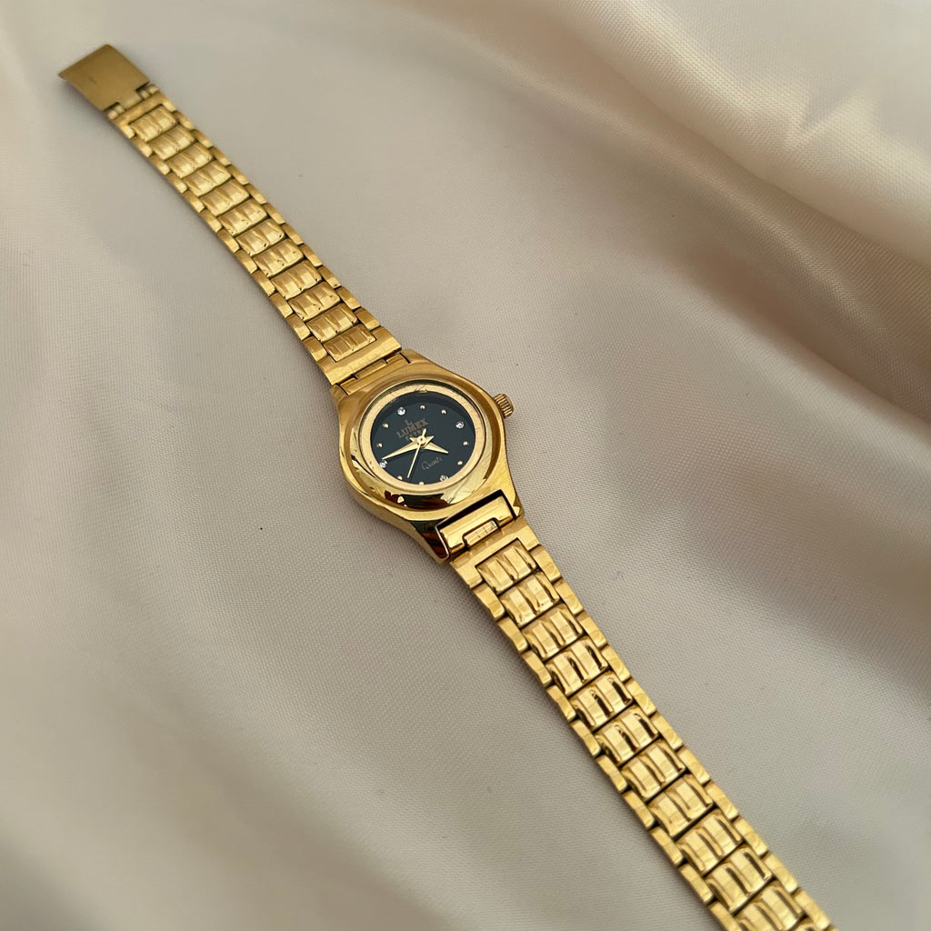 Round gold wristwatch with minimal dial and metallic strap on satin fabric background.